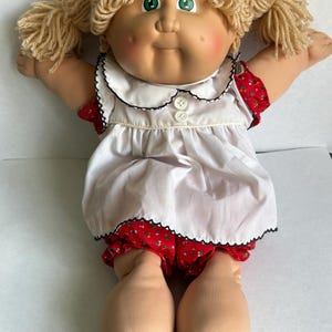 May include: A doll with light skin, green eyes, and blonde yarn hair styled in two pigtails. The doll wears a white short-sleeved dress with a black scalloped trim, two white buttons, and red floral bloomers with a small green leaf pattern.