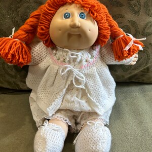 May include: A Cabbage Patch Kid doll with red yarn hair in pigtails, wearing a white crocheted cardigan and matching pants with a lace trim. The doll has blue eyes and a rosy complexion.