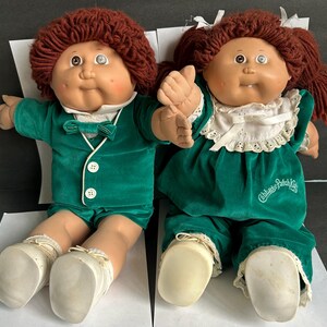 May include: Two Cabbage Patch Kids dolls with reddish-brown yarn hair and vinyl faces. The left doll wears a green velvet suit with a bow tie and white shoes. The right doll wears a green velvet dress with a white lace collar and white shoes.