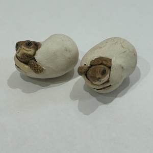 May include: Two small ceramic turtle figurines, each inside a cracked egg shell. The turtles are brown and have their heads poking out of the eggs. The eggs are white.