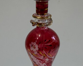 May include: A red glass bottle with a gold-trimmed stopper and a white floral design. The bottle has a round base and a tall, slender neck.