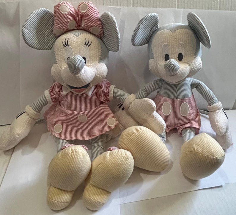 Puede incluir: Dos mu&ntilde;ecos de peluche de Mickey y Minnie Mouse. Mickey lleva pantalones cortos rojos, y Minnie lleva un vestido y un lazo a rayas rojas y blancas. Ambos mu&ntilde;ecos tienen cuerpos a rayas azules y blancas y manos y pies color crema.