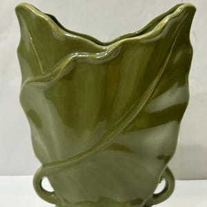May include: A green ceramic vase shaped like a leaf. The vase has two handles and a smooth, glossy finish.