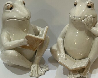 May include: Two white ceramic frog figurines sitting side-by-side, each holding a book. The frog on the left is reading a book with a brown cover, while the frog on the right is holding a book with a white cover that says "The Thirst" on it.