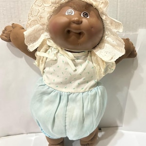 May include: A vintage doll with a dark complexion, wearing a cream-colored bonnet with lace trim and a light blue outfit. The doll has large, expressive eyes and outstretched arms, giving it a welcoming appearance. The doll's outfit includes a floral-printed top and a gathered skirt.