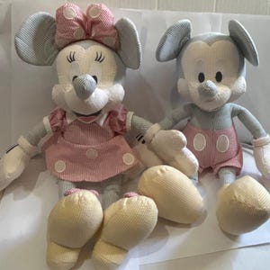 Puede incluir: Dos mu&ntilde;ecos de peluche de Mickey y Minnie Mouse. Mickey lleva pantalones cortos rojos con c&iacute;rculos blancos. Minnie lleva un vestido y un lazo rosas. Ambos mu&ntilde;ecos tienen cuerpos a rayas azul claro y blanco, guantes blancos y pies amarillos.