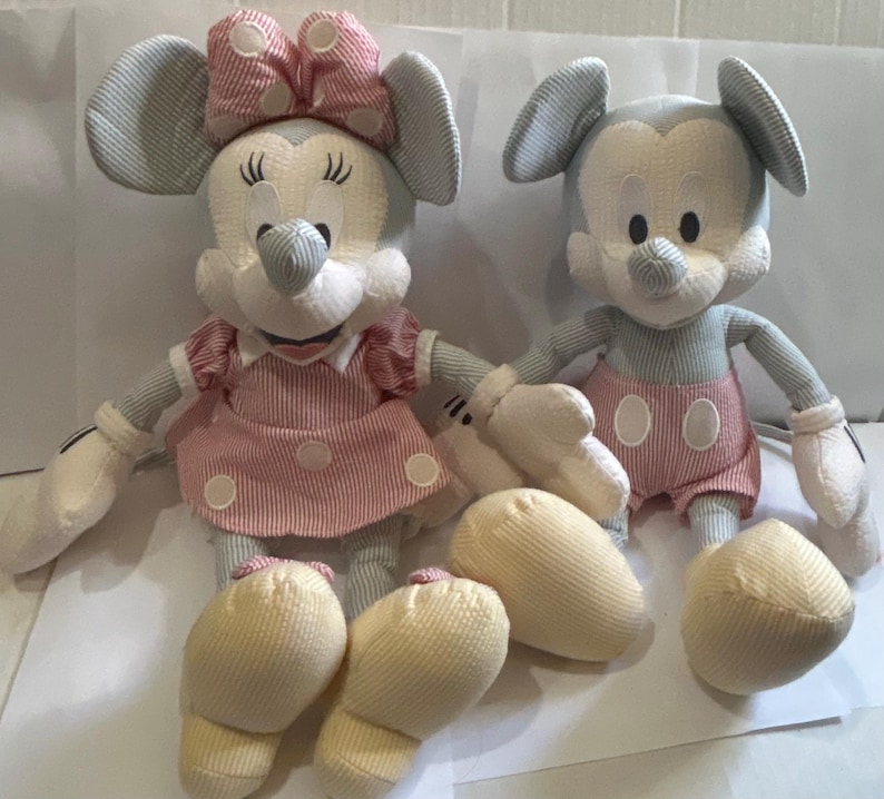 Puede incluir: Dos mu&ntilde;ecos de peluche de Mickey y Minnie Mouse. Mickey lleva pantalones cortos rojos con c&iacute;rculos blancos. Minnie lleva un vestido y un lazo rosas. Ambos mu&ntilde;ecos tienen cuerpos a rayas azul claro y blanco, guantes blancos y pies amarillos.