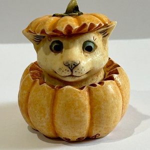 May include: A white ceramic cat figurine peeking out of a pumpkin. The cat has blue eyes and a small pink nose. The pumpkin is a light brown color with a green stem.