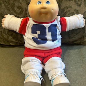 May include: A bald Cabbage Patch Kid doll wearing a red and white jersey with the number 31 on the front. The doll is sitting on a green couch.