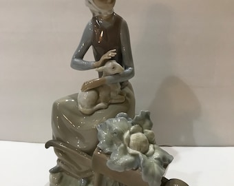 May include: A porcelain figurine of a girl in a white and blue polka dot headscarf holding a small white lamb. She is sitting in a wheelbarrow with a large cabbage.