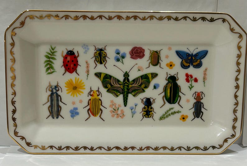 May include: A white rectangular ceramic tray with a gold rim and a decorative design of colorful insects and butterflies. The tray features a variety of insects, including ladybugs, beetles, and butterflies, all with intricate details.