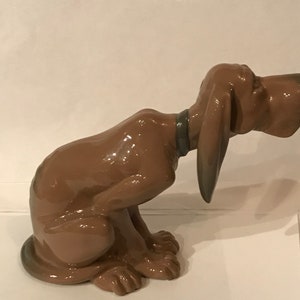 May include: A brown ceramic figurine of a bloodhound dog sitting with its head tilted down. The dog has a brown collar and is glazed with a shiny finish.