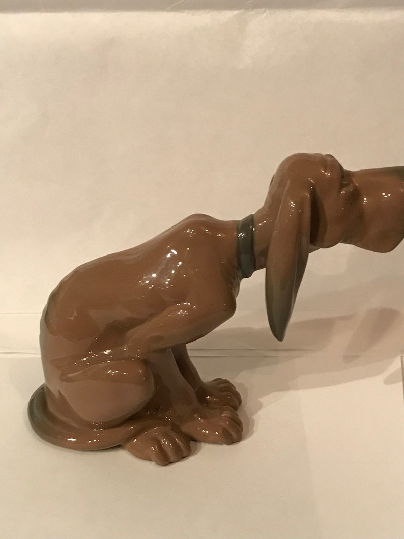 May include: A brown ceramic figurine of a bloodhound dog sitting with its head tilted down. The dog has a brown collar and is glazed with a shiny finish.