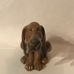 May include: A brown ceramic figurine of a dog with large floppy ears. The dog is sitting with its head tilted down and its nose pointing towards the ground.