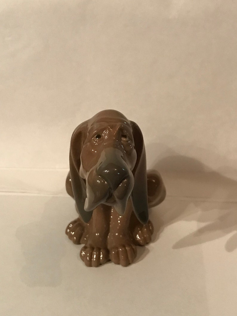May include: A brown ceramic figurine of a dog with large floppy ears. The dog is sitting with its head tilted down and its nose pointing towards the ground.
