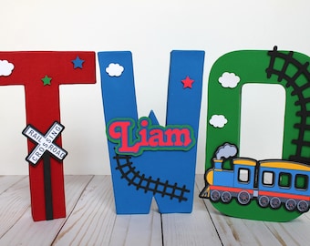May include: Three wooden letters spelling "TWO" decorated with a train theme. The letter "T" is red with a black and white railroad crossing sign, the letter "W" is blue with a black train track and the name "Liam" in red letters, and the letter "O" is green with a blue and yellow train with black wheels.