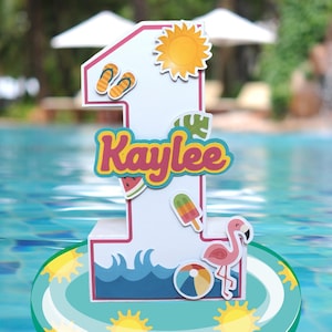 May include: A white and pink number one birthday decoration with summer-themed embellishments. The decoration includes a sun, flip-flops, a watermelon slice, an ice pop, a flamingo, and a beach ball. The name "Kaylee" is displayed in colorful letters.
