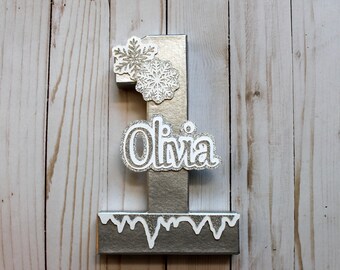 May include: Silver number one birthday decoration with the name "Olivia" in a glittery font. The number has snowflake accents and a dripping icicle design at the base. A festive decoration for a first birthday.