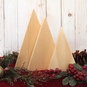 May include: Three wooden triangle shapes in varying sizes, arranged to resemble a row of Christmas trees. The trees are painted in a natural wood color and are sitting on a red surface with green and red holiday greenery.