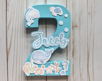 May include: A light blue wooden number two decorated with sea-themed cutouts. The number has a whale, a dolphin, a crab, a starfish, a seahorse, and a turtle. The name "Jacob" is written in white on the number.