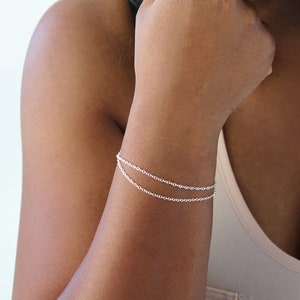 Puede incluir: Un brazalete de cadena de plata con dos cadenas delicadas. El brazalete se lleva en la muñeca izquierda de una persona con piel morena.