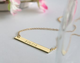 May include: Gold bar necklace with the word "SOLEIL" engraved on it.