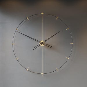 May include: A modern wall clock with a black frame and gold accents hangs on a gray wall. The clock's hands are black, and the hour markers are gold. Below the clock is a gray chaise lounge with pillows and a small side table.