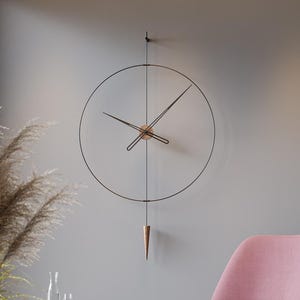 Könnte beinhalten: Ein modernes Wohnzimmer mit einer großen, minimalistischen Wanduhr mit schwarzem Rahmen und kupferfarbenen Akzenten. Ein rosa Sessel und ein passender Hocker stehen auf einem polierten Boden. Ein kleiner Beistelltisch aus Holz mit Glaskaraffe und Gläsern.