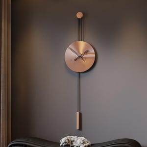 Op de afbeelding: Een moderne wandklok met een koperkleurige wijzerplaat en een slinger hangt boven een zwarte, gebogen bank. De wijzers van de klok zijn zwart. Een kleine, lichtgekleurde sculptuur rust op de bank. De muur is donkergrijs.
