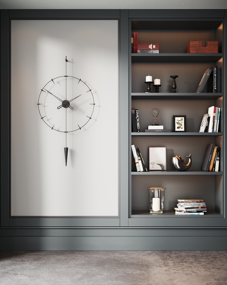 May include: A contemporary wall clock with a black metal frame and pendulum, set against a white wall. Adjacent is a built-in bookshelf, displaying books, candles, and ornaments. The colour palette is neutral, with dark grey accents throughout the room.