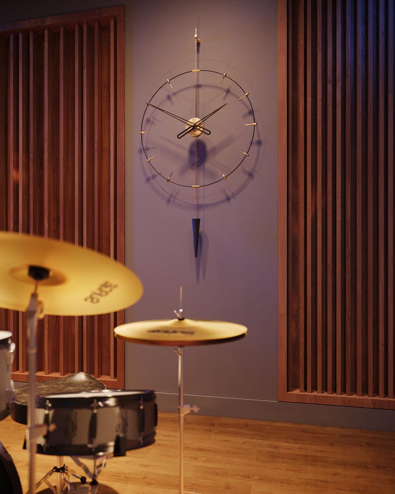 May include: A contemporary wall clock with a minimalist design. It has a gold-coloured centre and hands, with black lines indicating the hours. A dark blue, cone-shaped pendulum hangs below. The background includes wooden panelling and a drum kit.