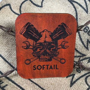 May include: A square, orange leather coaster with a black and white illustration of a skull wearing goggles and crossed wrenches. The skull is surrounded by two engine pistons. The text "SOFTTAIL" is printed below the skull.