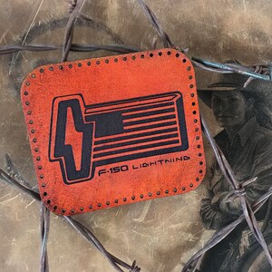 May include: Orange leather patch with a black and white graphic of a Ford F-150 Lightning truck. The text "F-150 LIGHTNING" is printed below the graphic.