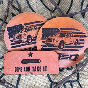 May include: Leather coasters and a small rectangular piece with the words "COME AND TAKE IT". The coasters feature a truck design with an American flag background. The rectangular piece has a star and a cannon design.