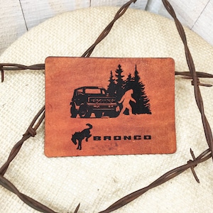 May include: A brown leather patch with a black silhouette of a Ford Bronco, a Bigfoot, and a horse. The text "BRONCO" is printed below the image.
