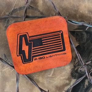 May include: A brown leather patch with a black graphic of a truck bed with an American flag design. The text "F-150 LIGHTNING" is printed below the graphic.