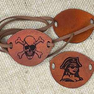 May include: Two brown leather pirate eye patches. One patch features a skull and crossbones design. The other patch features a pirate woman with an eyepatch and a hat.