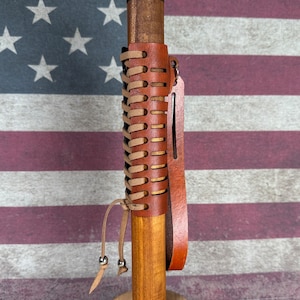 May include: A brown leather wrapped wooden walking stick with a tan leather strap and a brown leather tassel. The stick is standing upright against a red, white, and blue American flag.