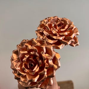 May include: Two metallic copper-coloured roses with layered, ruffled petals. The flowers feature a shiny, reflective surface with intricate folds that mimic the natural appearance of blooming rose blossoms, captured in a bright, clear light.