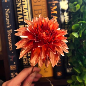 May include: A metallic copper-coloured flower sculpture with numerous pointed, layered petals radiating from a central point. The flower has a reflective, shiny surface and is attached to a thin, dark stem, showcasing intricate craftsmanship.