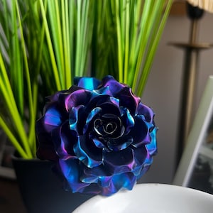 May include: A metallic rose sculpture featuring iridescent petals that shift between deep shades of purple, blue, and teal. The flower has a multi-layered, blooming structure with curved, reflective edges that catch the light, creating a vibrant colour gradient across the surface of the petals.
