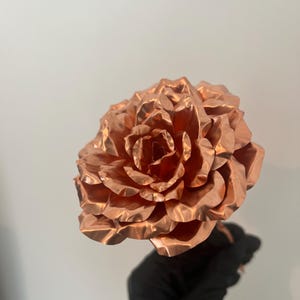 May include: A large, copper-colored metal rose with detailed petal construction. The rose is held by a gloved hand against a plain background. The metallic finish gives the flower a reflective quality.