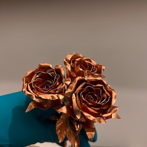 May include: A cluster of three metallic copper roses with intricately layered petals and detailed leaves. The flowers exhibit a polished, reflective surface with warm, reddish-brown tones, showcasing a realistic floral structure crafted from metal.