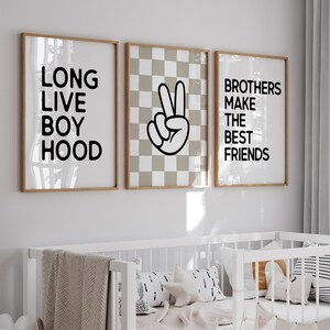 Puede incluir: Una cuna blanca con una manta beige y almohadas decorativas está frente a tres impresiones enmarcadas. Las impresiones tienen las frases "LONG LIVE BOY HOOD", un signo de paz y "BROTHERS MAKE THE BEST FRIENDS" en texto negro.