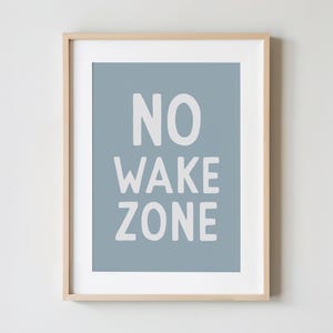 May include: A framed print with the words "NO WAKE ZONE" in white block letters on a light blue background. The print is in a light wood frame and is displayed against a white wall.