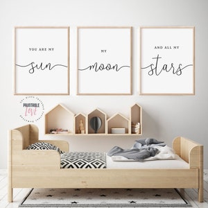 May include: Three framed prints with white backgrounds and black calligraphy. The prints read "You are my sun", "My moon", and "And all my stars". The prints are hanging on a white wall above a wooden bed with a black and white patterned blanket.
