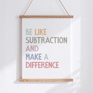 May include: A white poster with the text "BE LIKE SUBTRACTION AND MAKE A DIFFERENCE" in pastel colours. The poster is held by wooden bars at the top and bottom, suspended by a natural rope.