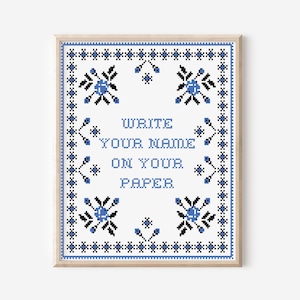 May include: A framed cross-stitch artwork with a light wood frame. The design features a white background with blue and black floral and geometric patterns. The text "WRITE YOUR NAME ON YOUR PAPER" is centered in blue.