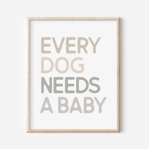 Op de afbeelding: Een ingelijste print met de tekst "EVERY DOG NEEDS A BABY" in verschillende tinten grijs en beige. De woorden zijn gecentreerd op een witte achtergrond, en de lijst is licht van kleur.