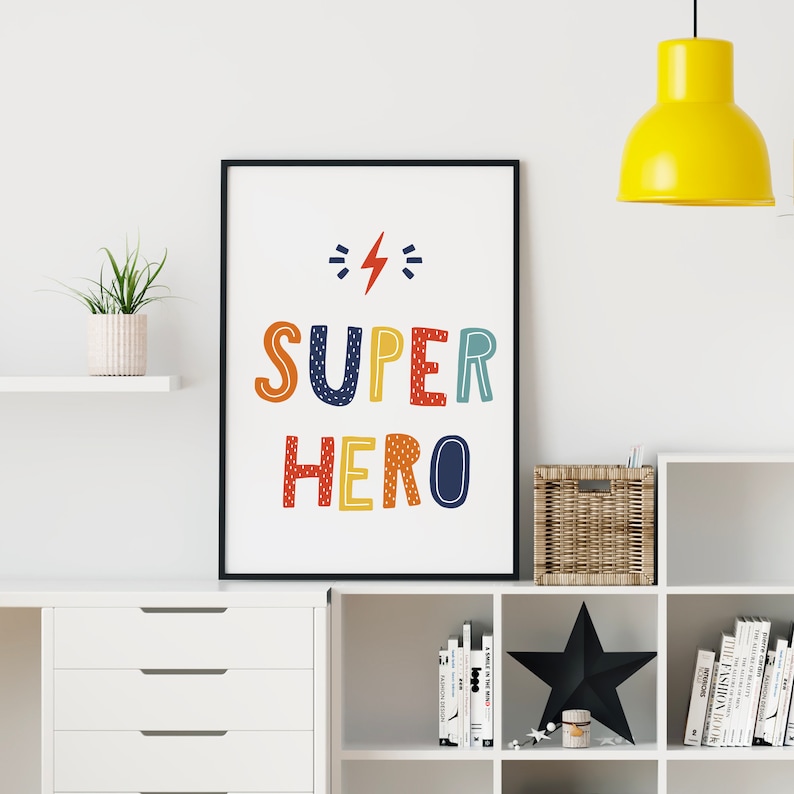 以下が含まれることがあります： 白い背景と黒いフレームの額装されたプリント。プリントには、"SUPER"の文字の上に稲妻が描かれた、カラフルでスタイリッシュな文字で"SUPER HERO"という文字が書かれています。